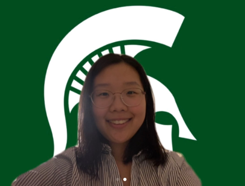Girl wearing glasses smiling at camera in front of a Zoom MSU Sparty head background. 
