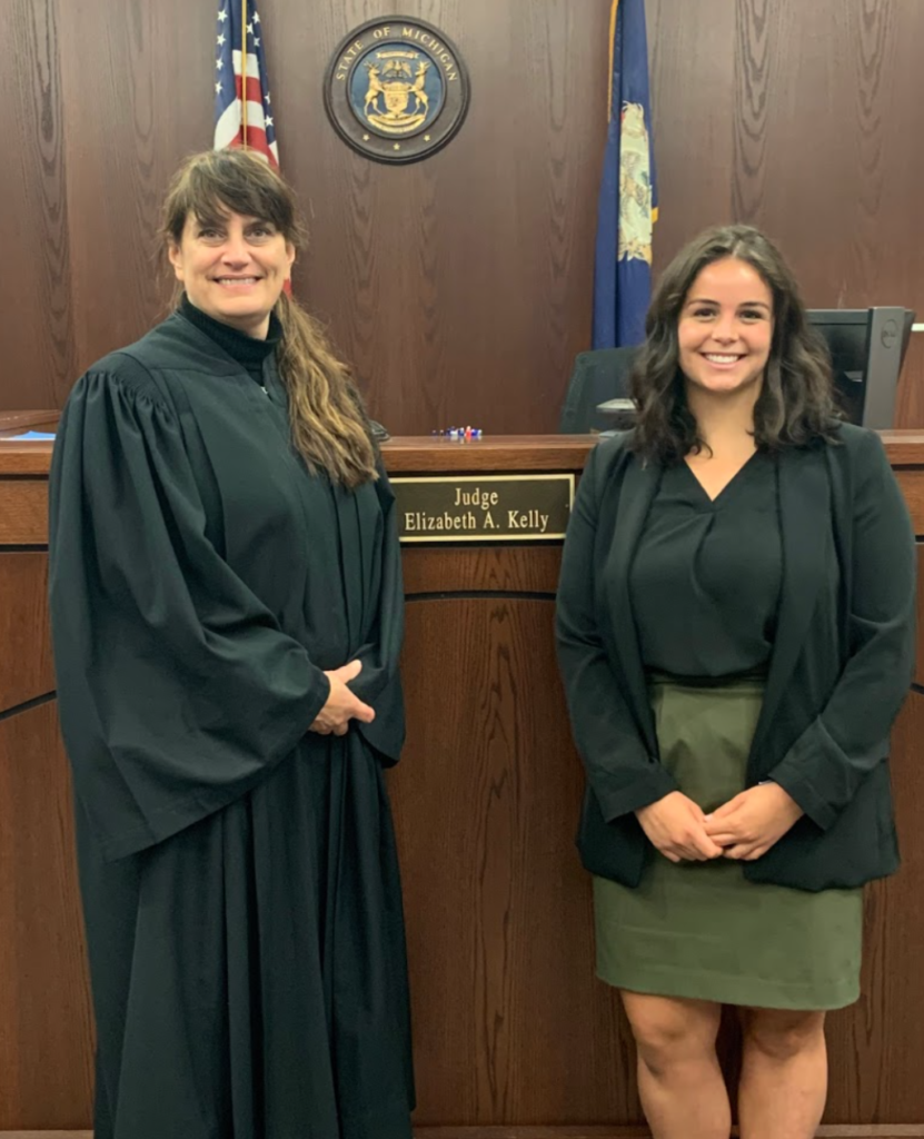 Two women, one in judge robes and one in formal wear smiling at camera in front of a judge desk. 