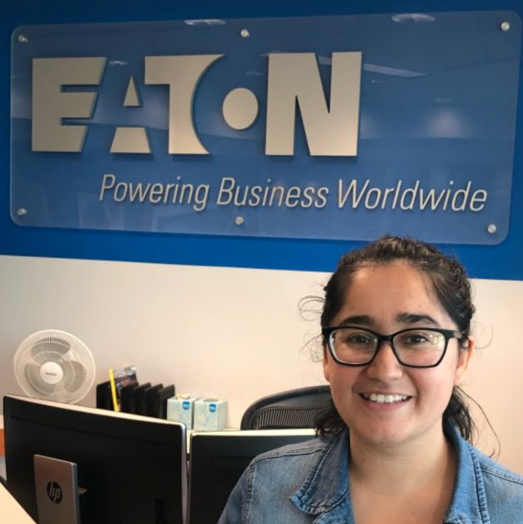 Woman in glasses smiling in front of an EATON Powering Business Worldwide logo sign. 