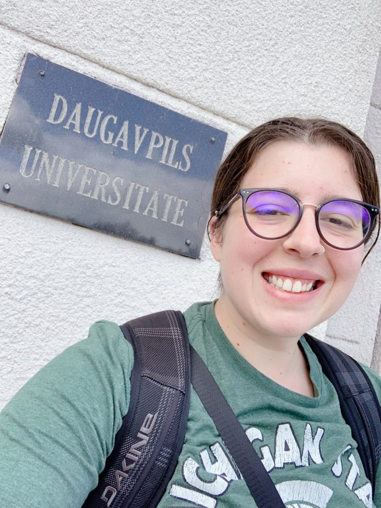 A young woman standing in front of a "Daugavpils Universitate" gray sign. 